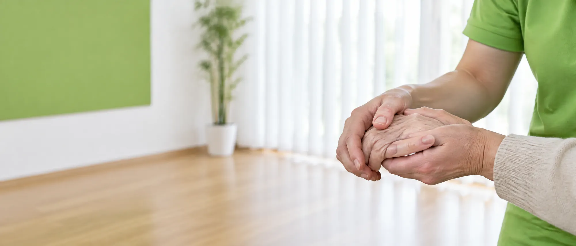 Therapeutenhände im grünen Poloshirt halten behutsam die Hand einer älteren Person, im Hintergrund ein grünes Wandfeld und Bambus